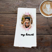 A white towel with a photo of a baby and the text "my heart" printed on it, placed on a wooden surface next to a cup of coffee.