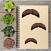 a notebook with stickers on it, surrounded by four potted plants on a wooden surface.
