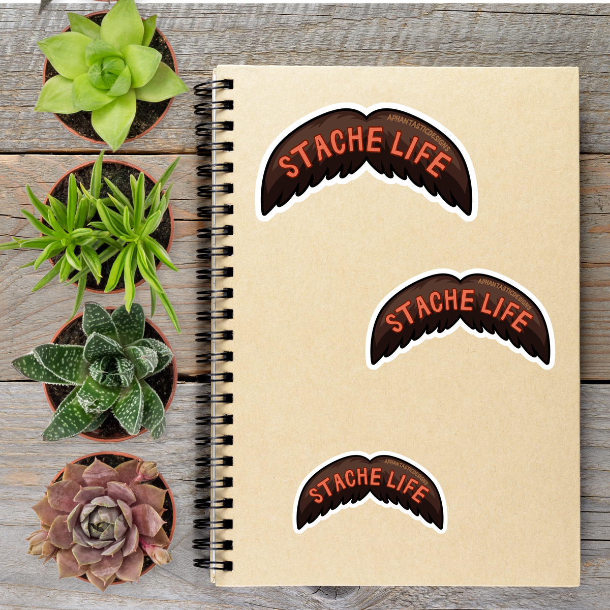 a notebook with stickers on it, surrounded by four potted plants on a wooden surface.