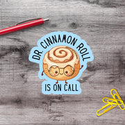 A sticker with a cinnamon roll character and the text "Dr. Cinnamon Roll is on Call" is placed on a wooden surface next to a red pen and yellow paper clips.
