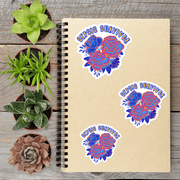 a spiral-bound notebook with three stickers on it, surrounded by four potted succulent plants on a wooden surface.