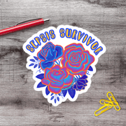 A sticker with a floral design and the text "SILIPS SURVIVOR" is placed on a wooden surface next to a red pen and yellow paper clips.